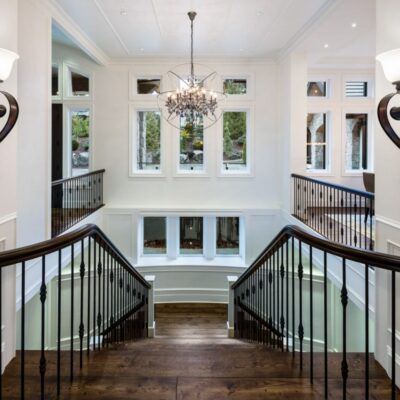 View from the top of a grand staircase with wall sconces, showcasing soaring ceilings and large windows in a custom-designed Langley home.