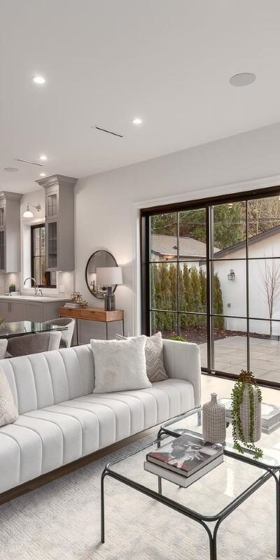 A bright living room with large picture windows revealing the backyard. A grey modern renovated kitchen behind the sofa in an open concept living space.