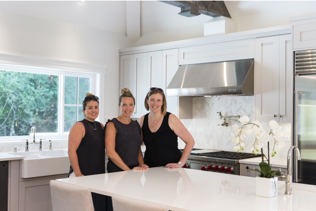 Three happy women stand behind a large white kitchen island in a freshly renovated space.