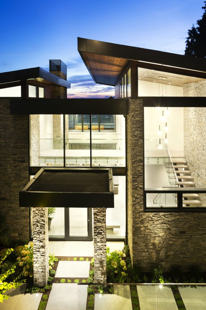 A warmly lit home exterior featuring a glass entry door, picture windows, and a vaulted rooftop with additional windows enhancing natural light and architectural charm.