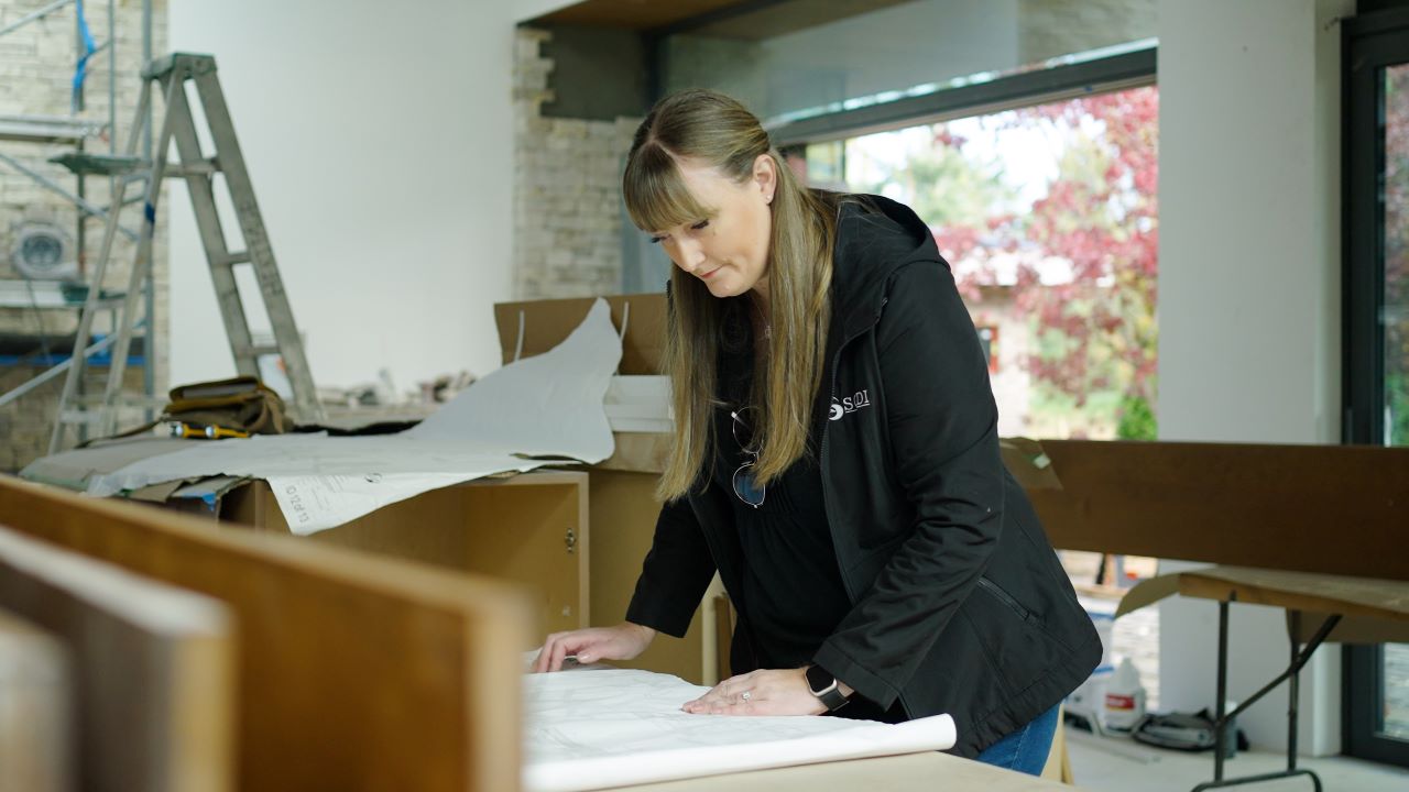 Designer Sarah Gallop looks over plans for an upcoming design in an office surrounded by large papers and designs.
