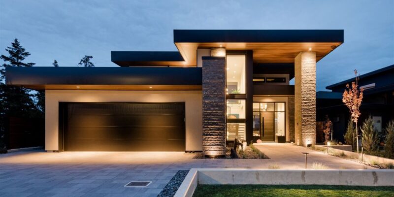 Front view of a striking modern hillside home with clean lines, flat rooflines, and warm accent lighting highlighting its neutral exterior and architectural presence.