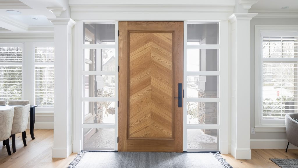 Bright, symmetrical entryway with a solid wood front door in a chevron pattern, flanked by glass sidelights, white trim, and light hardwood flooring in a renovated South Surrey home.