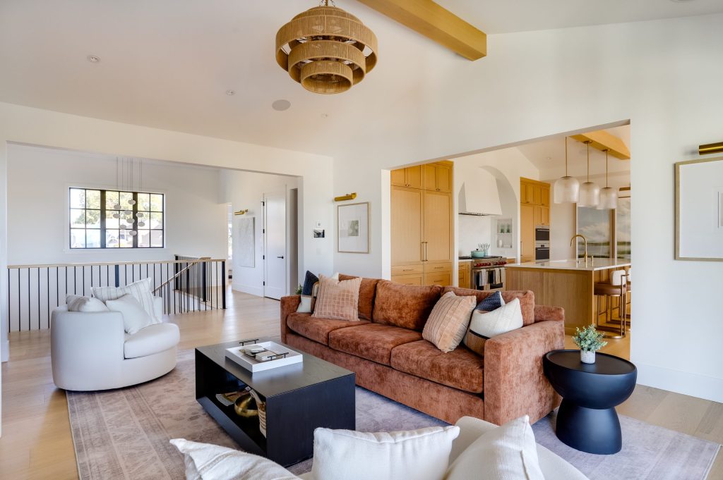 Bright open-concept living room and kitchen with warm wood cabinetry, vaulted ceiling with exposed beam, terracotta sofa, neutral furnishings, and modern lighting in a contemporary BC home by Sarah Gallop Design